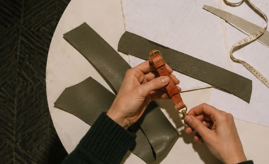 Hands fastening a hand-crafted terracotta leather collar on a workbench of woven straps